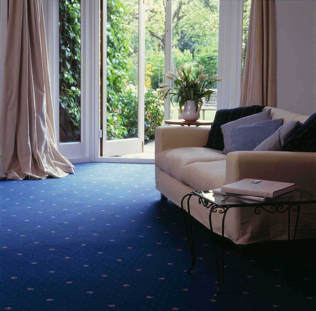 Cremesofa und Beistelltisch in einem Zimmer mit blauem Teppichboden und offener Tür zum Garten