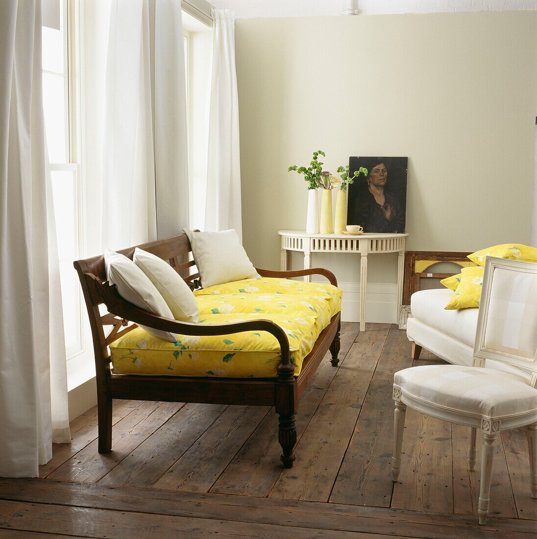 Yellow upholstered daybed at windows of room with exposed floorboards and portrait on console table