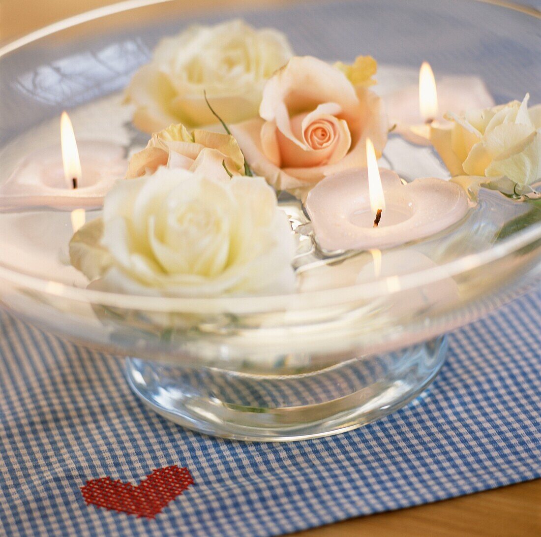 Rosen schwimmen in einer Schale mit Wasser und brennenden Kerzen auf kariertem Stoff mit gesticktem Herz