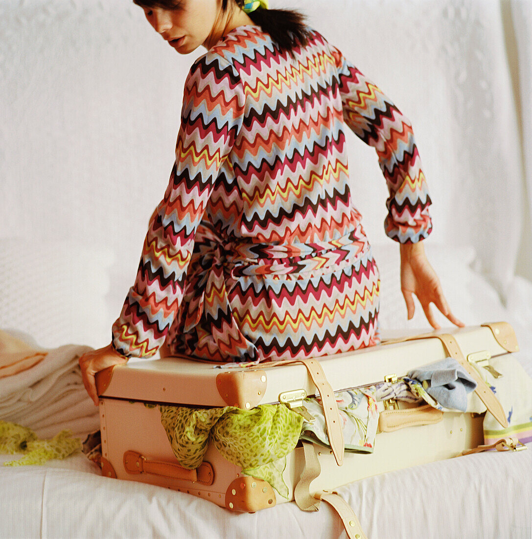 Frau im gestreiften Kleid sitzt auf einem überquellenden Koffer