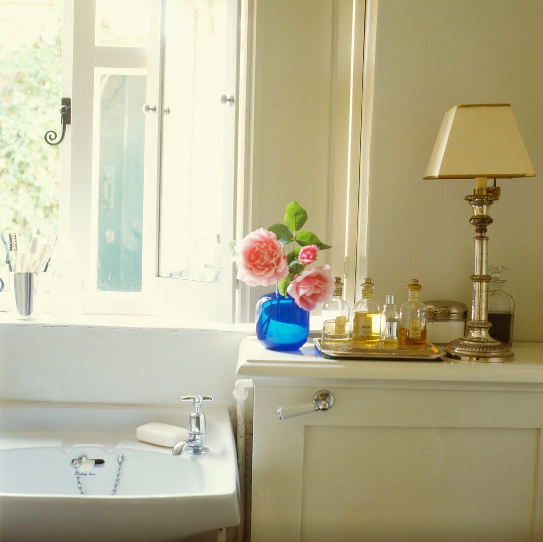 Waschbecken im Badezimmer unter dem Fenster mit Schnittblumen und einem Tablett mit Parfüm und Lampe