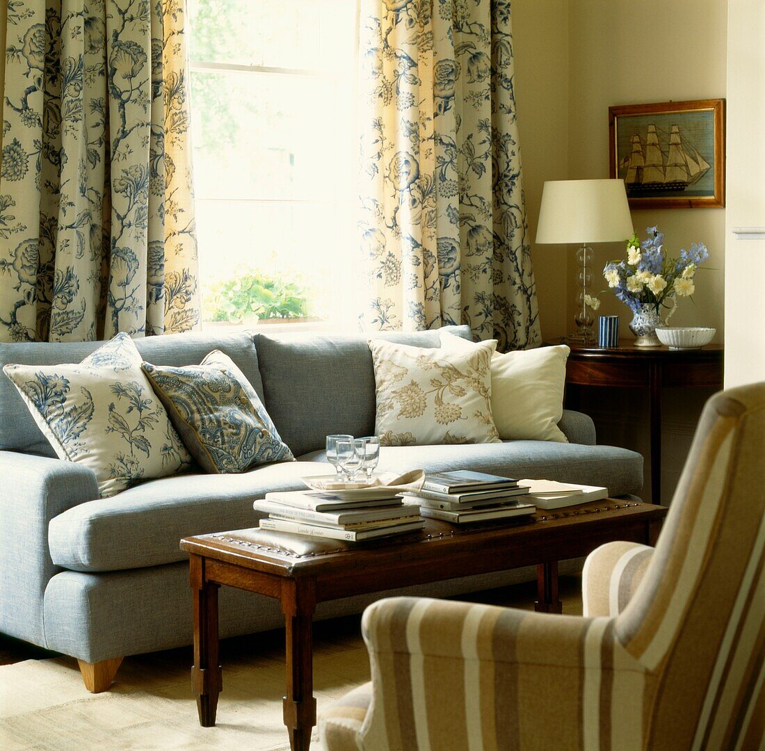 Hellblaues Sofa mit Kissen, dazu passende Vorhänge und gestreifter Sessel auf der gegenüberliegenden Seite eines Couchtisches mit Büchern