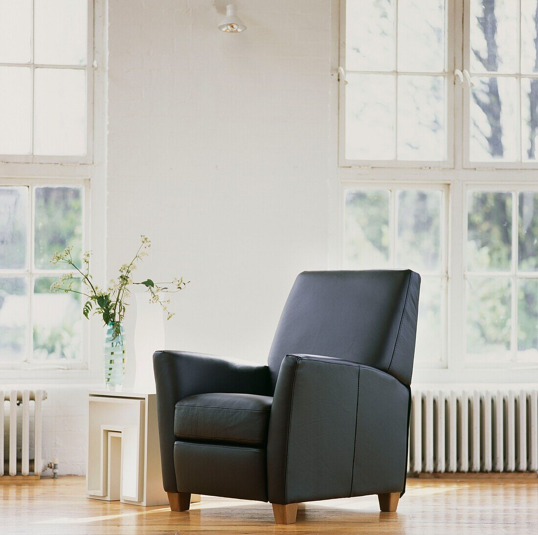 Black leather armchair and flower arrangement on nest of tables in white room with sunlit sash windows