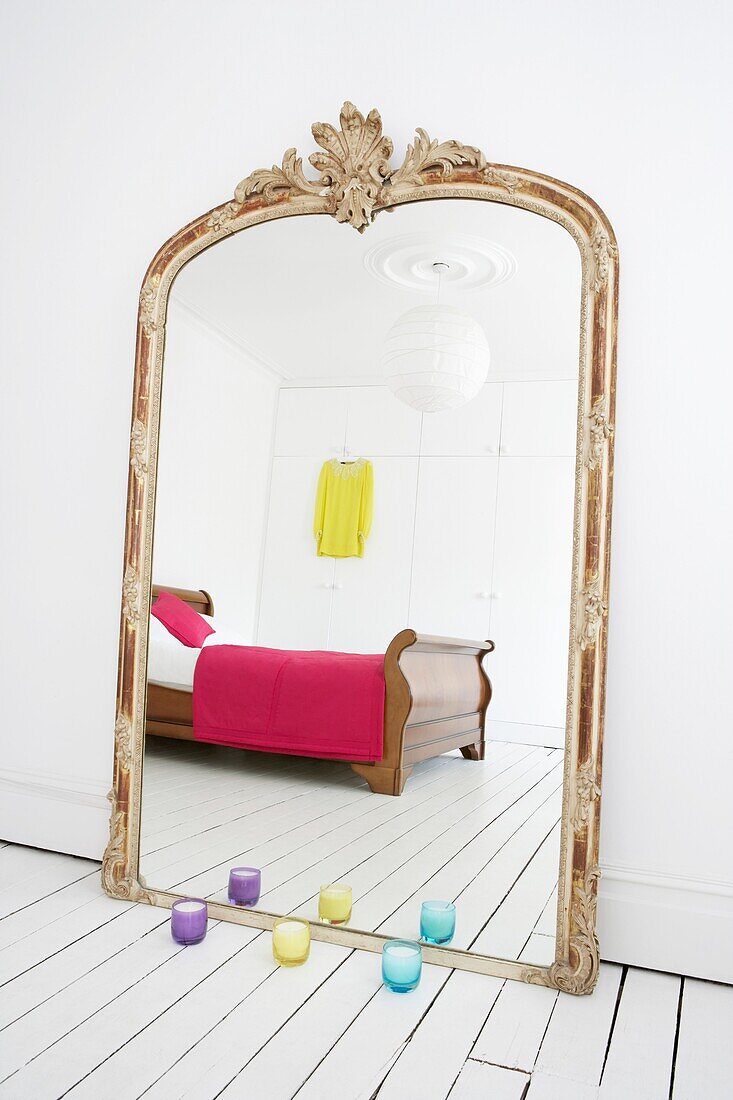 Übergroßer verschnörkelter Vintage-Spiegel in einem ganz in Weiß gehaltenen Schlafzimmer mit Schlittenbett und Schränken, die sich im Spiegel und in bunten Accessoires spiegeln