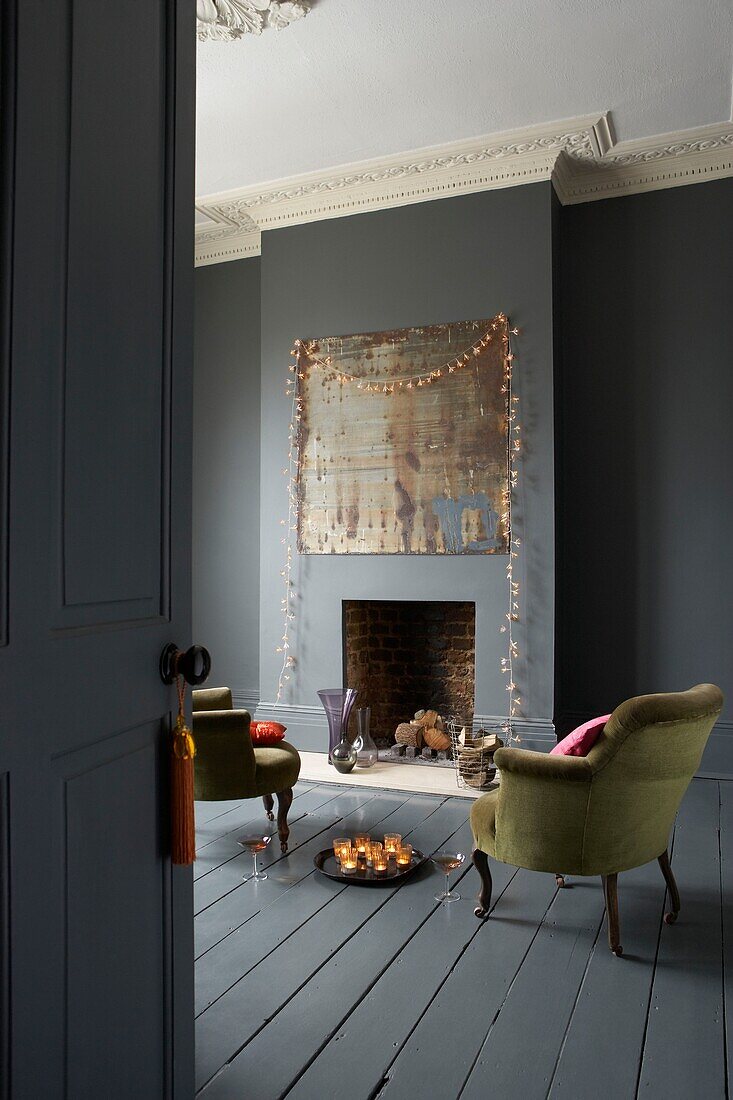 Tray of candles and champagne on floor in dark grey painted living room with fairylights draped around the fireplace 