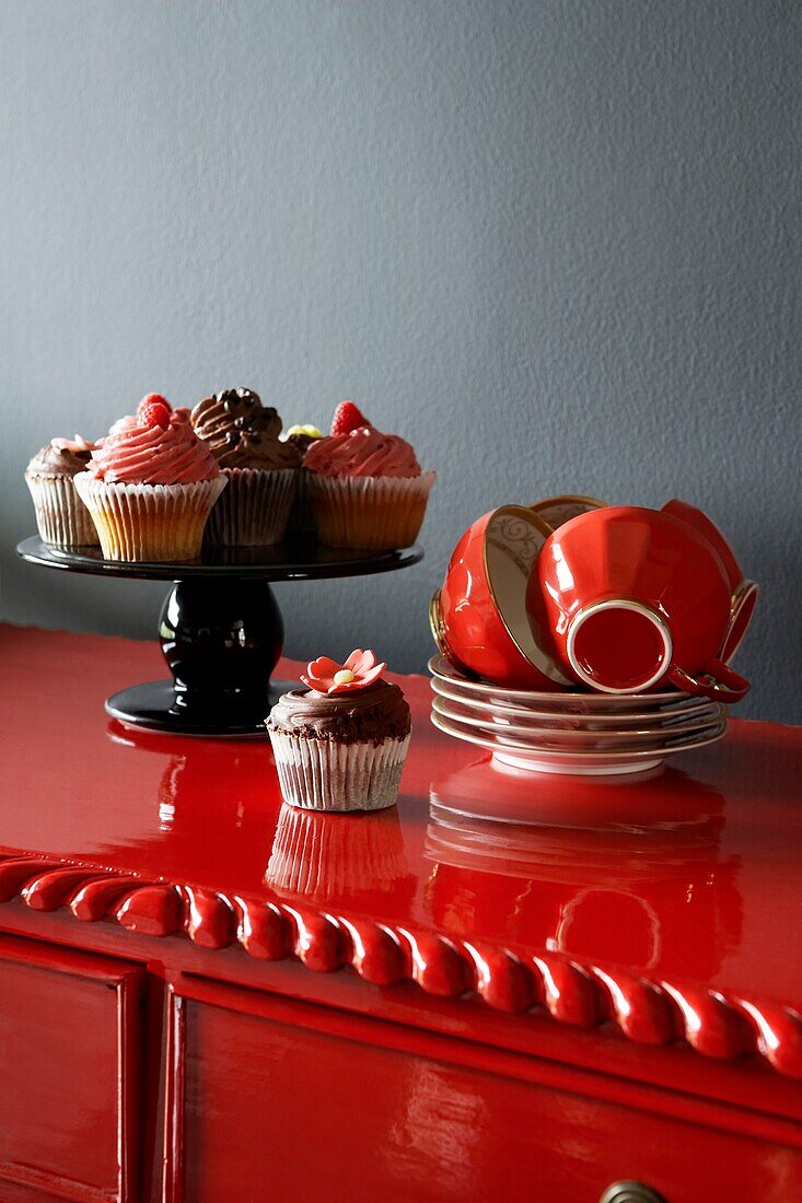 Leuchtend rot gestrichene Anrichte vor einer grauen Wand mit roten Tassen und Untertassen, die neben einem mit Tassenkuchen gefüllten Kuchenstand gestapelt sind
