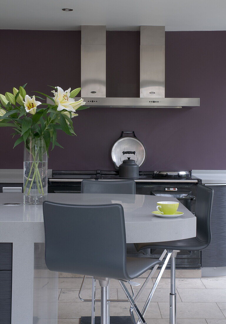 Grey bar stools with cut lilies on breakfast bar of purple kitchen in contemporary Haywards Heath home,  West Sussex,  England,  UK