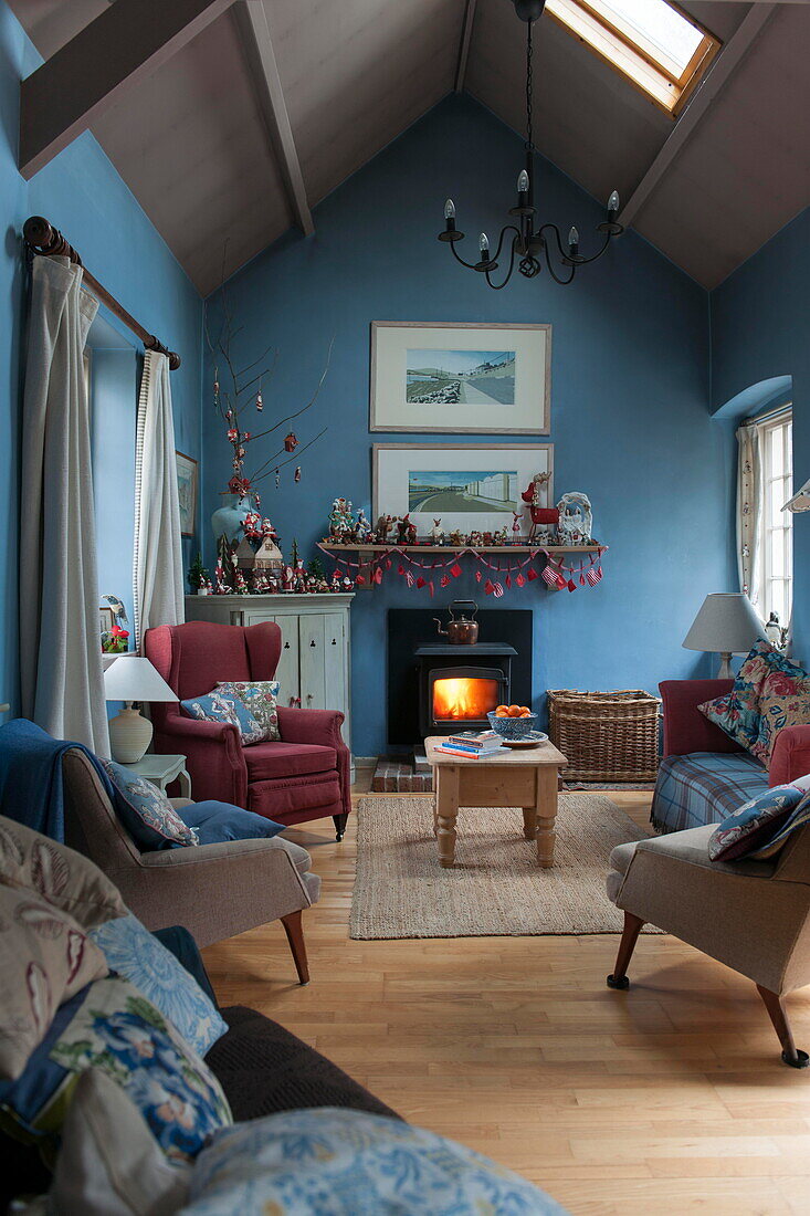 Sessel und Sofa mit beleuchtetem Holzofen im blauen Wohnzimmer eines Bauernhauses in Tiverton, Devon, Großbritannien
