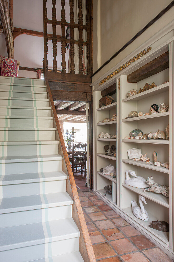 Collection of ornaments on hallway shelving with painted stairs in Suffolk home  England  UK