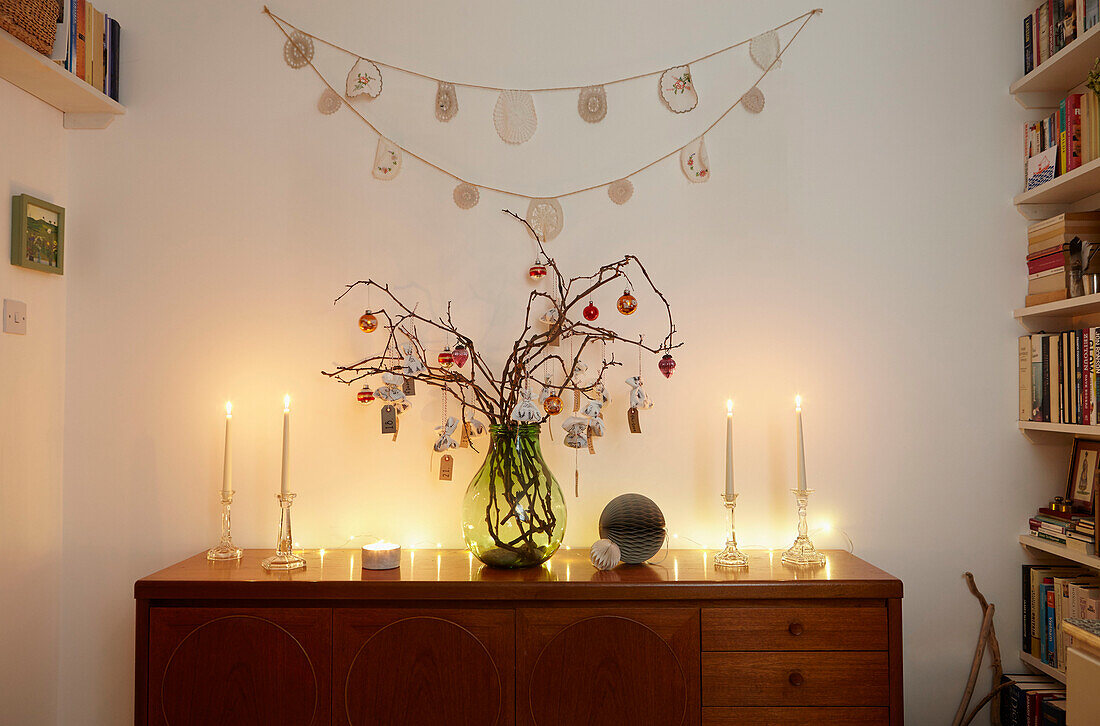 Weihnachtskugeln auf Zweig-Arrangement mit brennenden Kerzen auf Sideboard in Londoner Wohnung UK