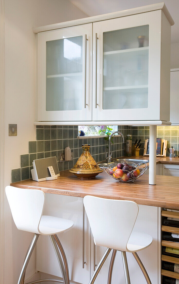 Glass fronted fitted units above wooden kitchen worktop with bar stools in New Malden home, Surrey, England, UK