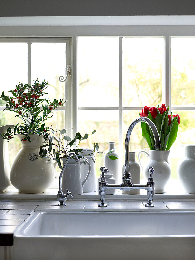 Detail der Küchenspüle und des Fensters mit Flaschen, Krügen und Gläsern, gefüllt mit roten Tulpen und Blättern