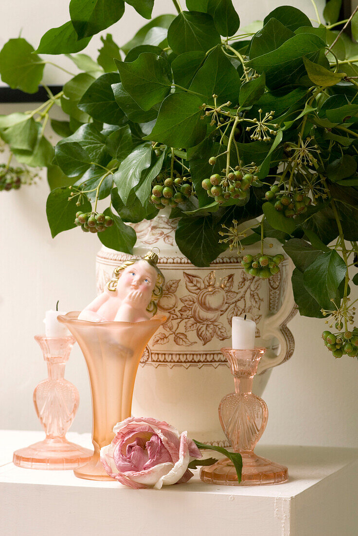 Porzellanvase mit Efeublättern neben rosafarbenen Glaskerzenhaltern und Vase mit Vintage-Kugel und frischem Rosenkopf auf dem Kaminsims