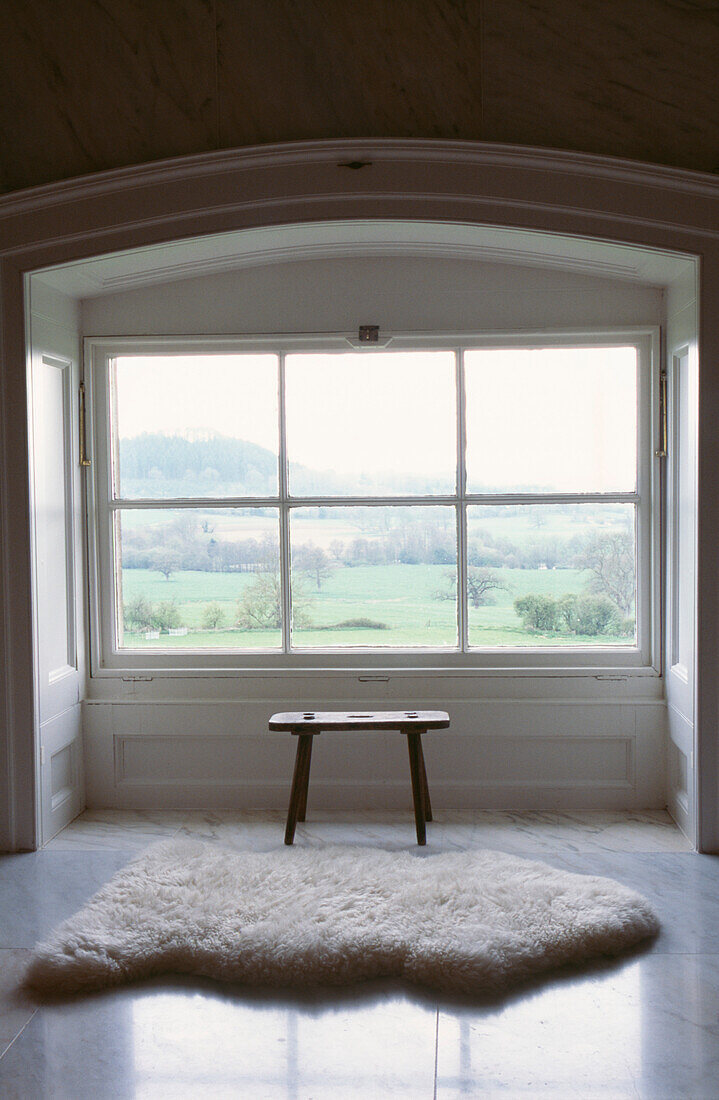 Schaffellteppich und Holzhocker vor einem Fenster ohne Vorhang