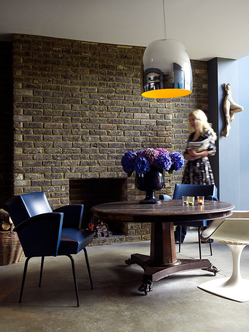 Frau, die durch einen Essbereich mit Ledersessel und Vase mit blauen und violetten Hortensien auf einem runden Tisch vor einem gemauerten Schornstein mit Kamin geht