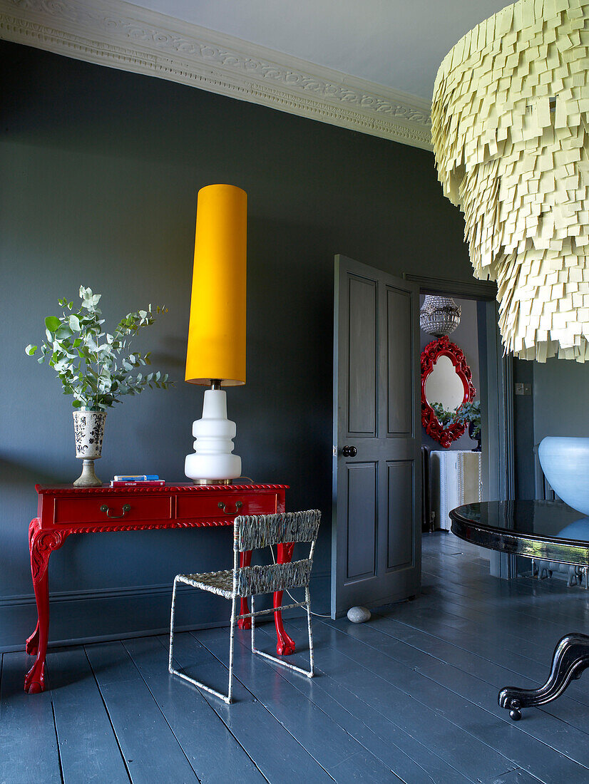 Mittelblauer, grau dekorierter Raum mit leuchtend rot bemaltem Konsolentisch und gelber Lampe und Kronleuchter im Zwischengeschoss
