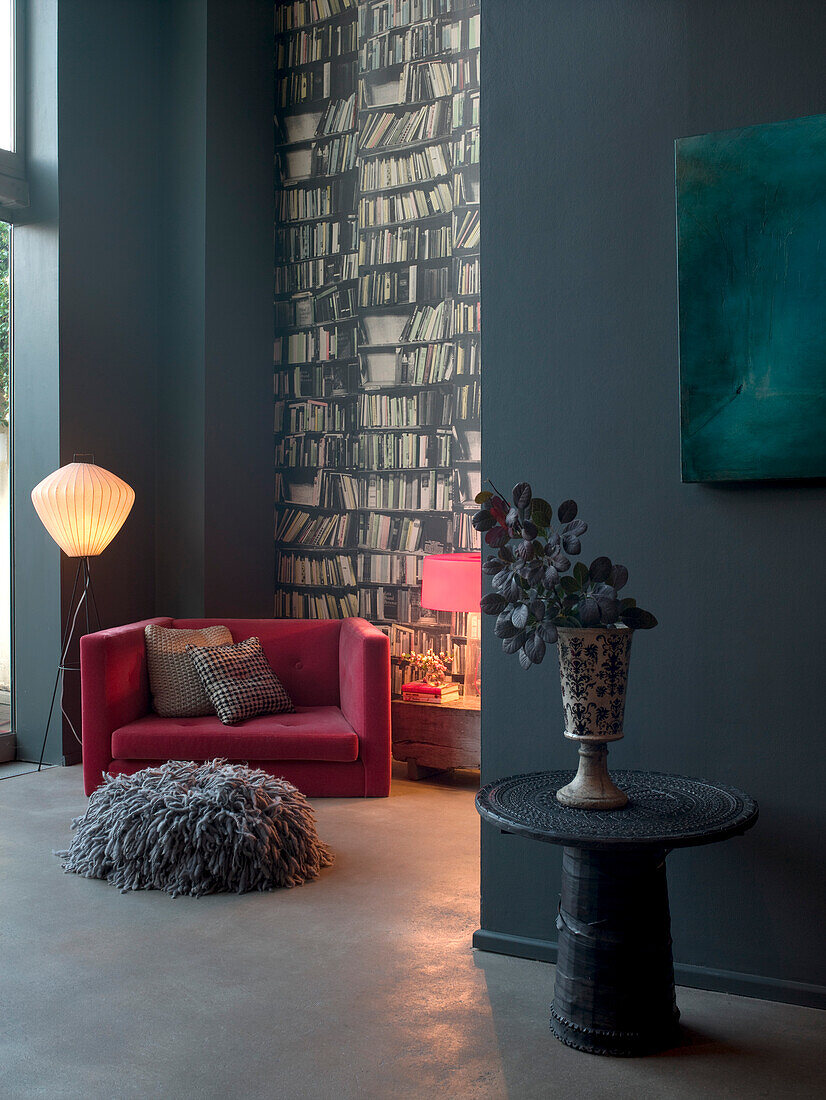 Rosafarbener Sessel an einer mit Büchertapete dekorierten Wand mit grauem Bodenkissen und Beistelltisch mit Vase