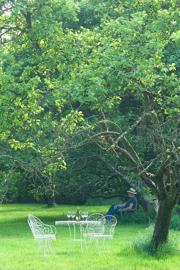 Mann mit Sonnenhut sitzt im Obstgarten mit Weinflaschen auf dem Tisch