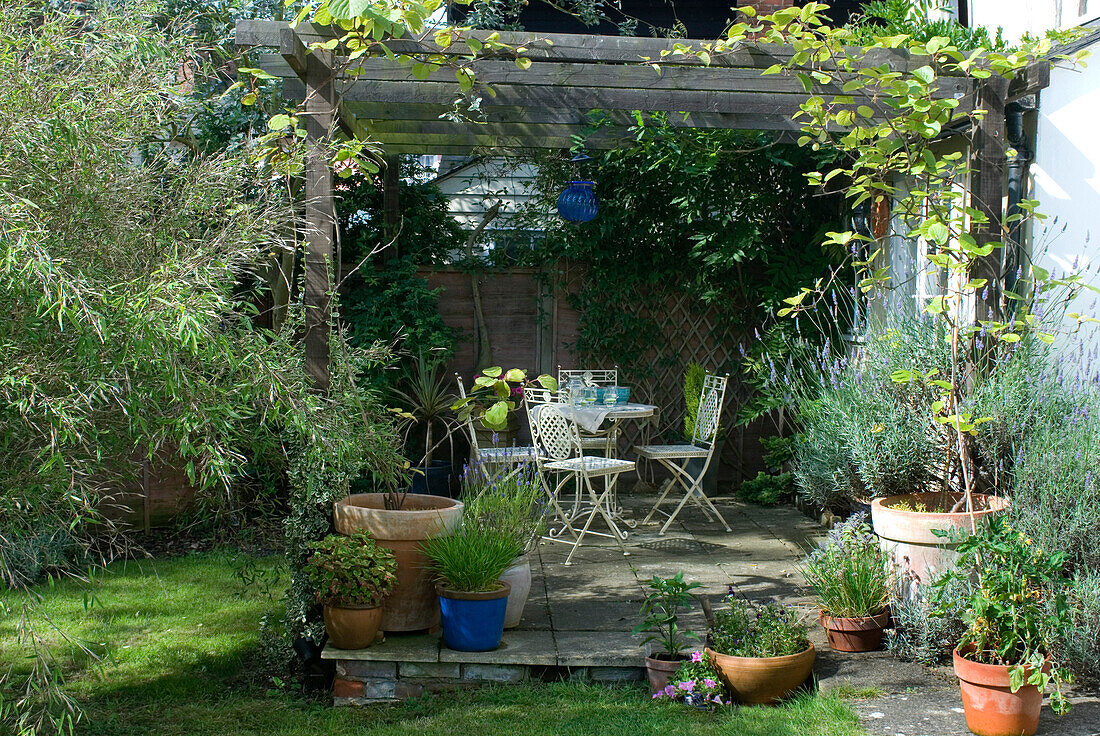 Terrassenbereich im hinteren Garten mit weißen Ziermetall-Gartenmöbeln und einer blauen Glaslaterne, die an einer rustikalen Laube hängt