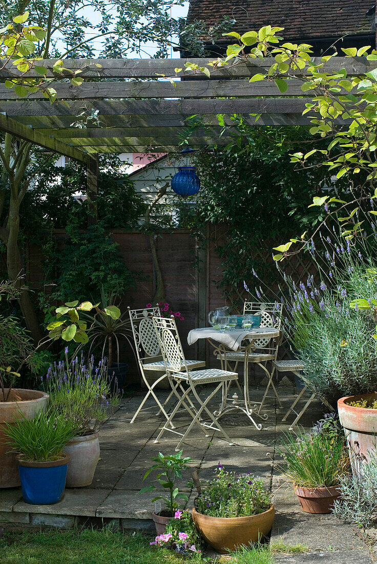 Terrassenbereich im hinteren Garten mit weißen Ziermetall-Gartenmöbeln und einer blauen Glaslaterne, die an einer rustikalen Laube hängt