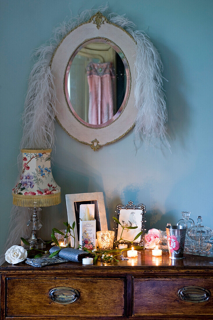 Weiße Federboa hängt über einem Spiegel über einem hölzernen Beistelltisch mit Bilderrahmen und brennenden Kerzen