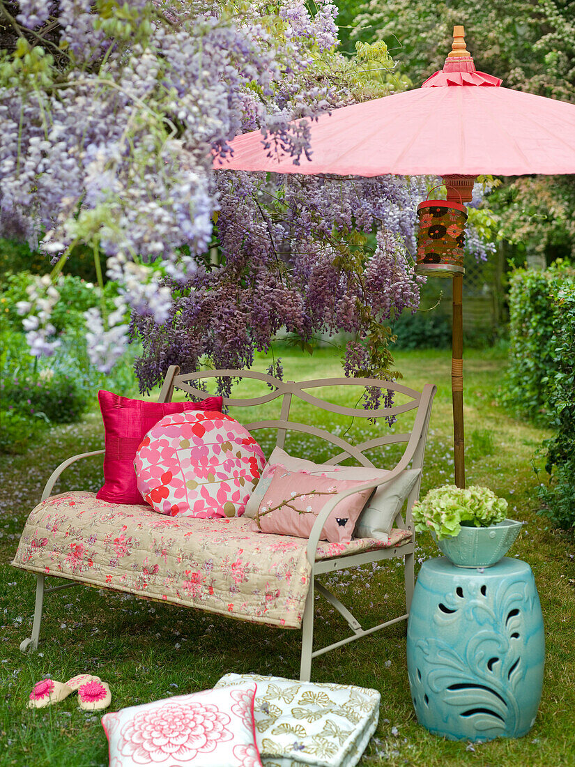 Sitzbank mit Kissen und Überwürfen in einem Sommergarten mit lila Glyzinien und einem Sonnenschirm