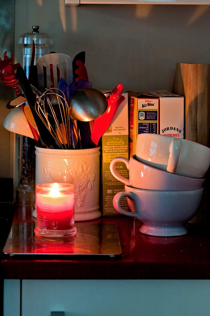 Utensilienhalter und brennende Kerze mit gestapelten Teetassen in der Küche eines Londoner Hauses UK