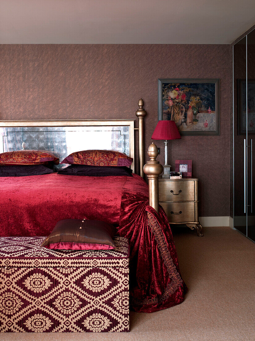 Red velvet bed cover on gold bed with patterned blanket box in bedroom of London apartment England UK