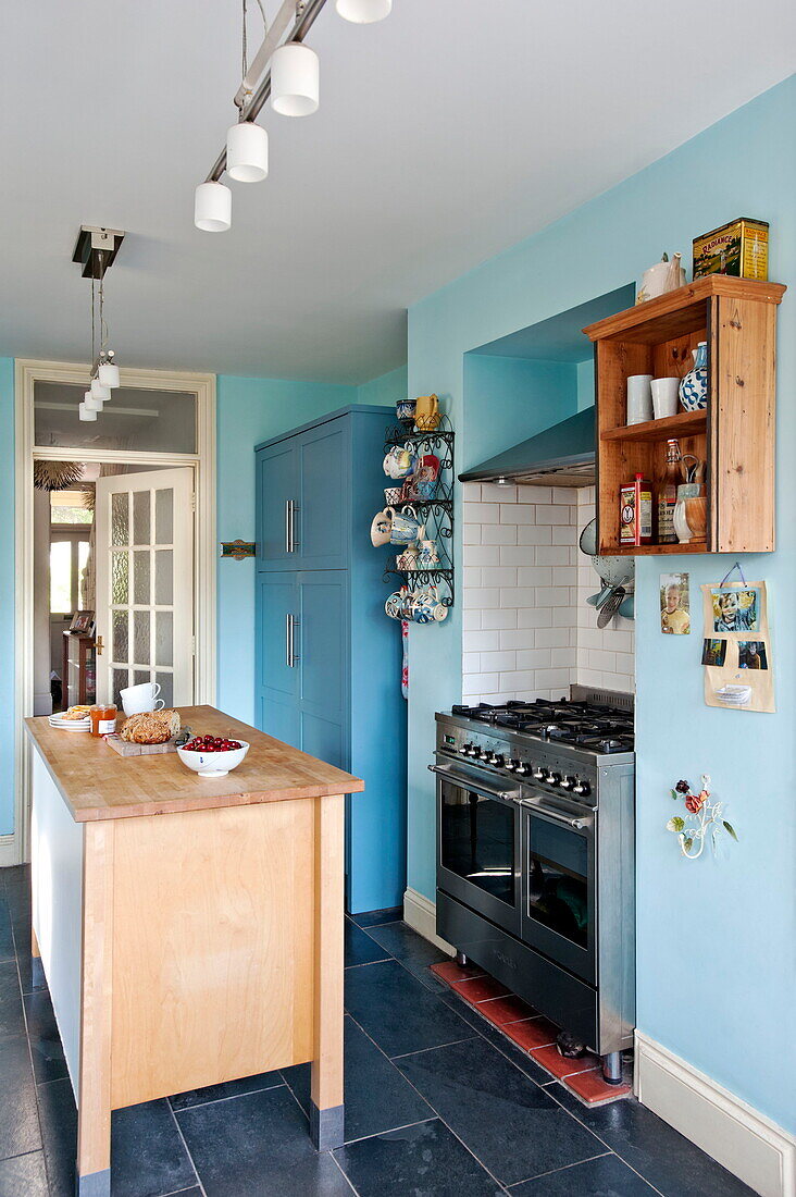 Holzinsel mit wandmontierten Regalen in blauer Küche eines Einfamilienhauses in Bovey Tracey, Devon, England, UK