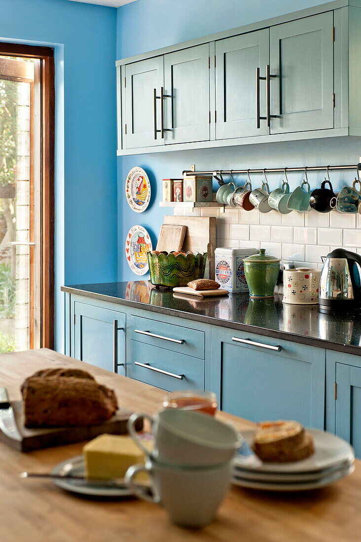 Tassen, Wasserkocher, Geschirr und Brot auf der Arbeitsplatte in der blauen Einbauküche eines Familienhauses in Bovey Tracey, Devon, England, UK