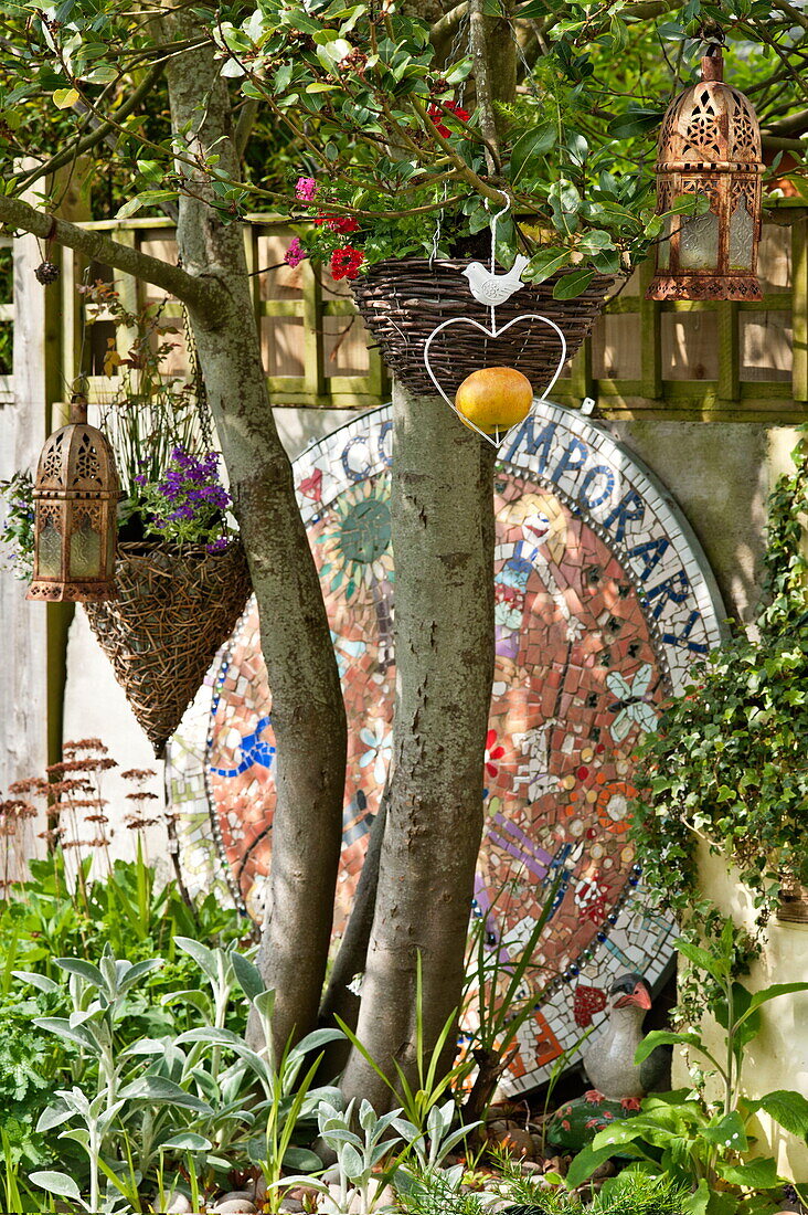 Gartenornamente hängen an einem Baum mit großem Mosaik im Garten von Bovey Tracey, Devon, England, UK