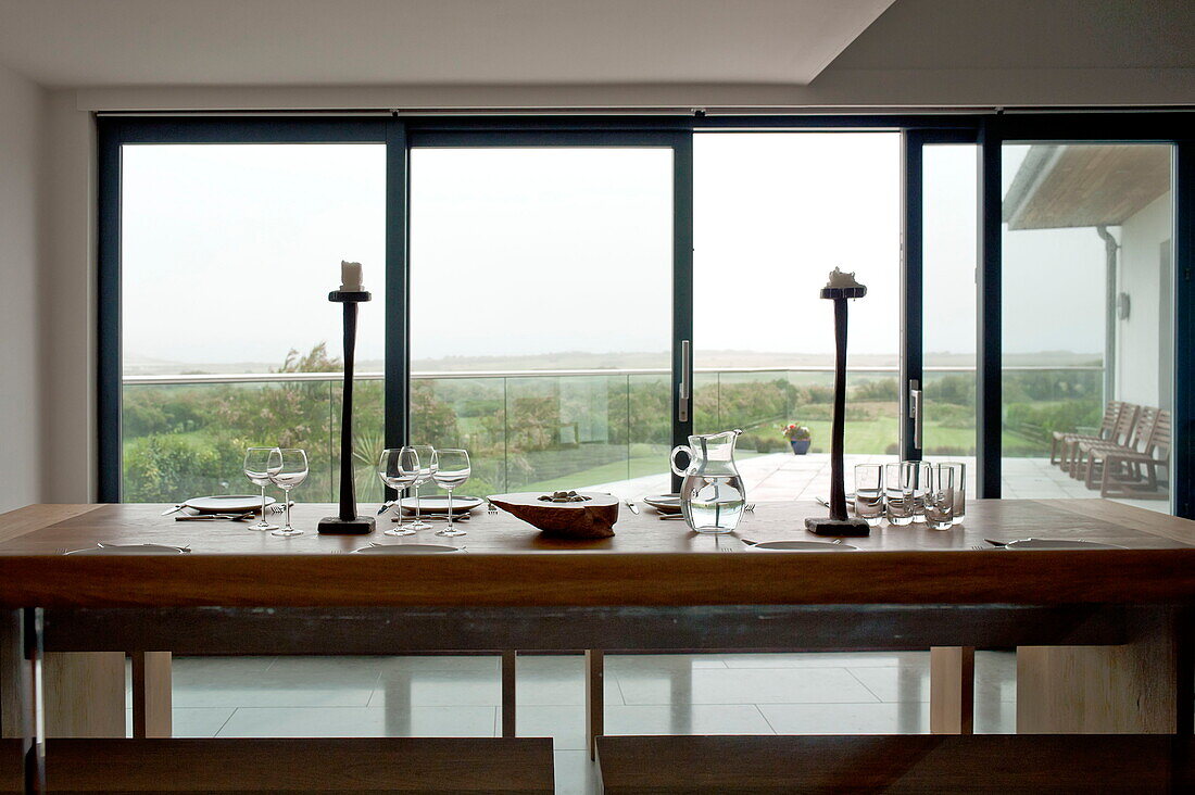 Wooden dining table with bench seats and view of countryside from contemporary detached home, Cornwall, England, UK