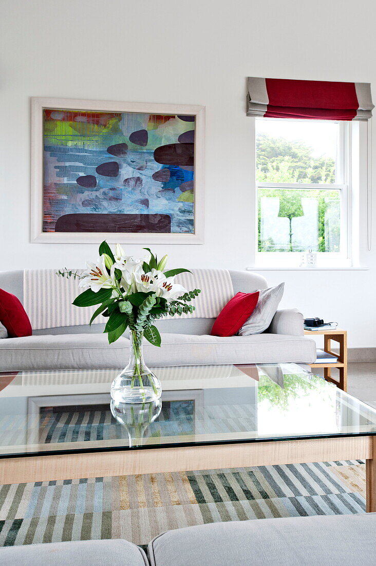 Lilien auf einem gläsernen Couchtisch mit Sofa und Kunstwerken in einem modernen Haus in Cornwall, England, Vereinigtes Königreich