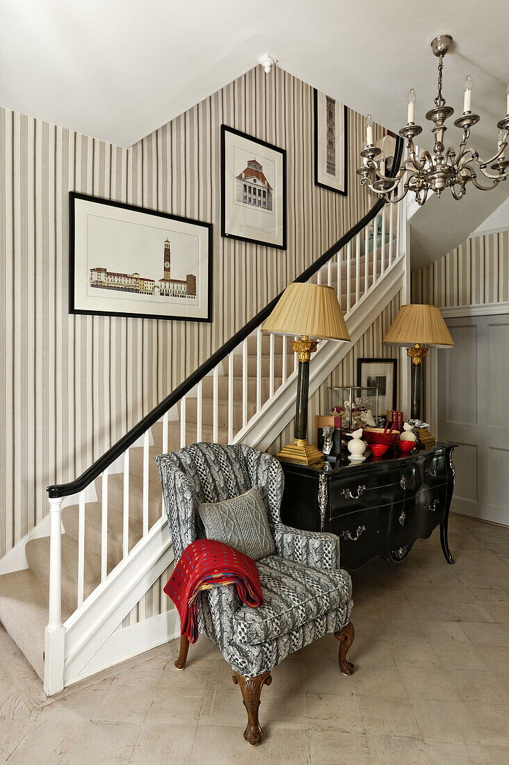 Grey upholstered armchair with black sideboard under stairs in entrance of Bury St Edmunds country home, Suffolk, England, UK
