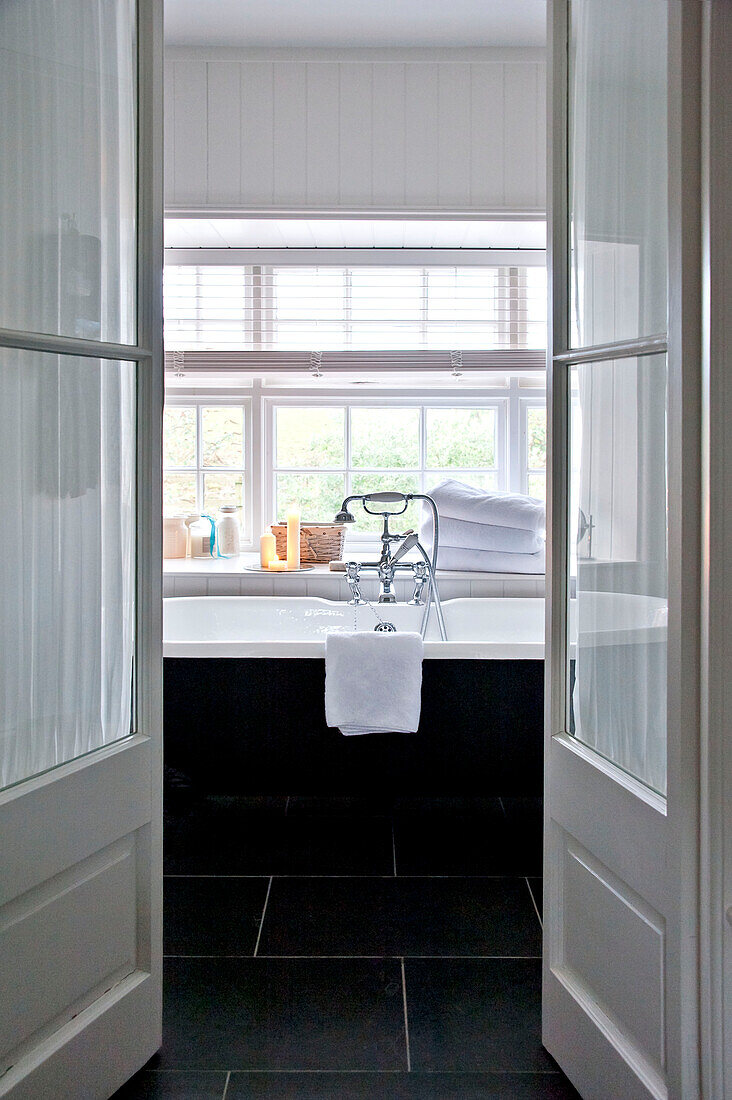 Blick auf eine freistehende Badewanne unter dem Fenster durch eine verglaste Doppeltür im Haus in Crantock, Cornwall, England UK
