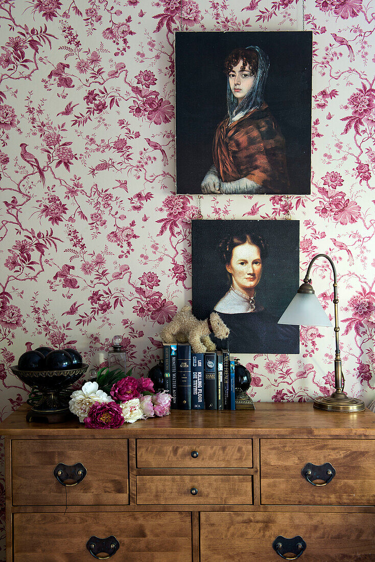 Kunstwerkporträts auf Blumentapete über einer Holztruhe mit Vintage-Lampe in einem Haus in Stamford, Lincolnshire, England, UK