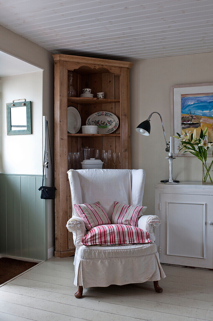 Striped cushions on slip-covered wingback armchair with wooden cabinet in corner of beach house Cornwall England UK