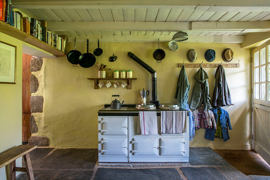 Bücher auf einem Regal mit Mänteln an Haken in einer Bauernhausküche mit weißem Gasherd in Helston, Cornwall, UK