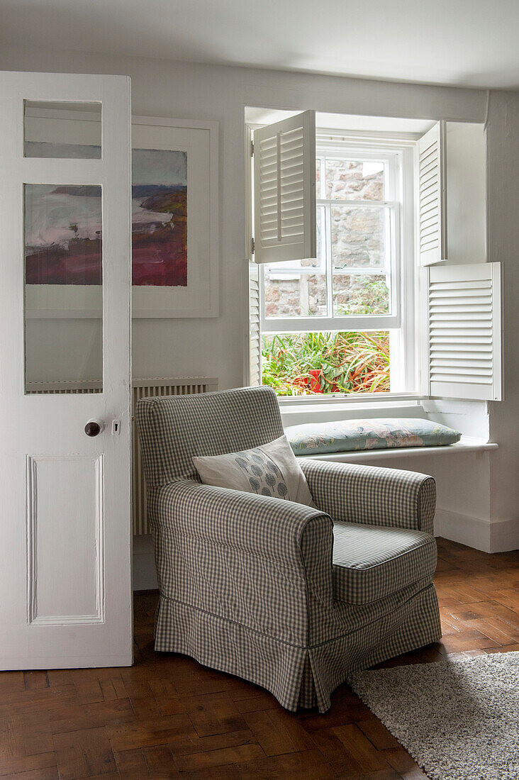 Gingham-Sessel am Sprossenfenster mit Fensterläden im Cottage-Wohnzimmer in Cornwall, UK