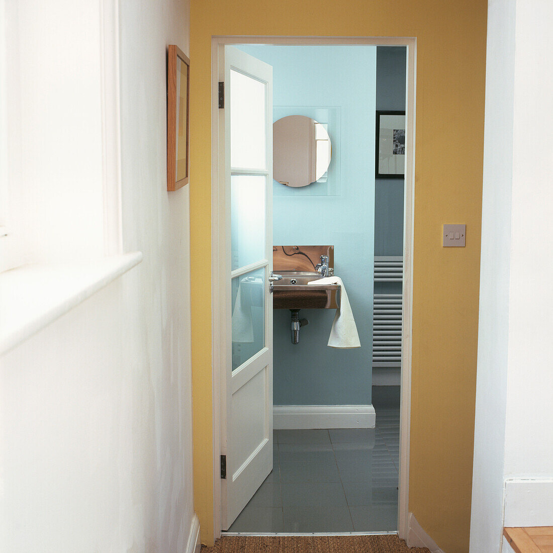 View of blue painted bathroom with metal basin through glass doorway in yellow painted hall