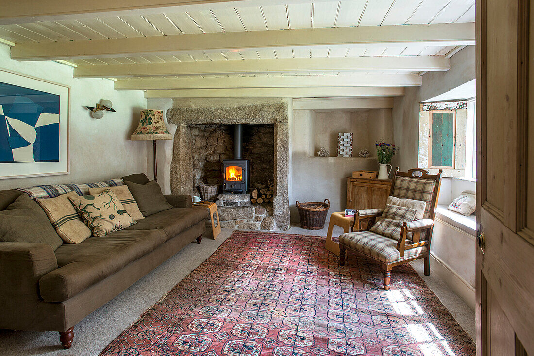 Braunes Sofa am Kamin mit beleuchtetem Holzofen im Wohnzimmer eines Bauernhauses in Helston, Cornwall UK