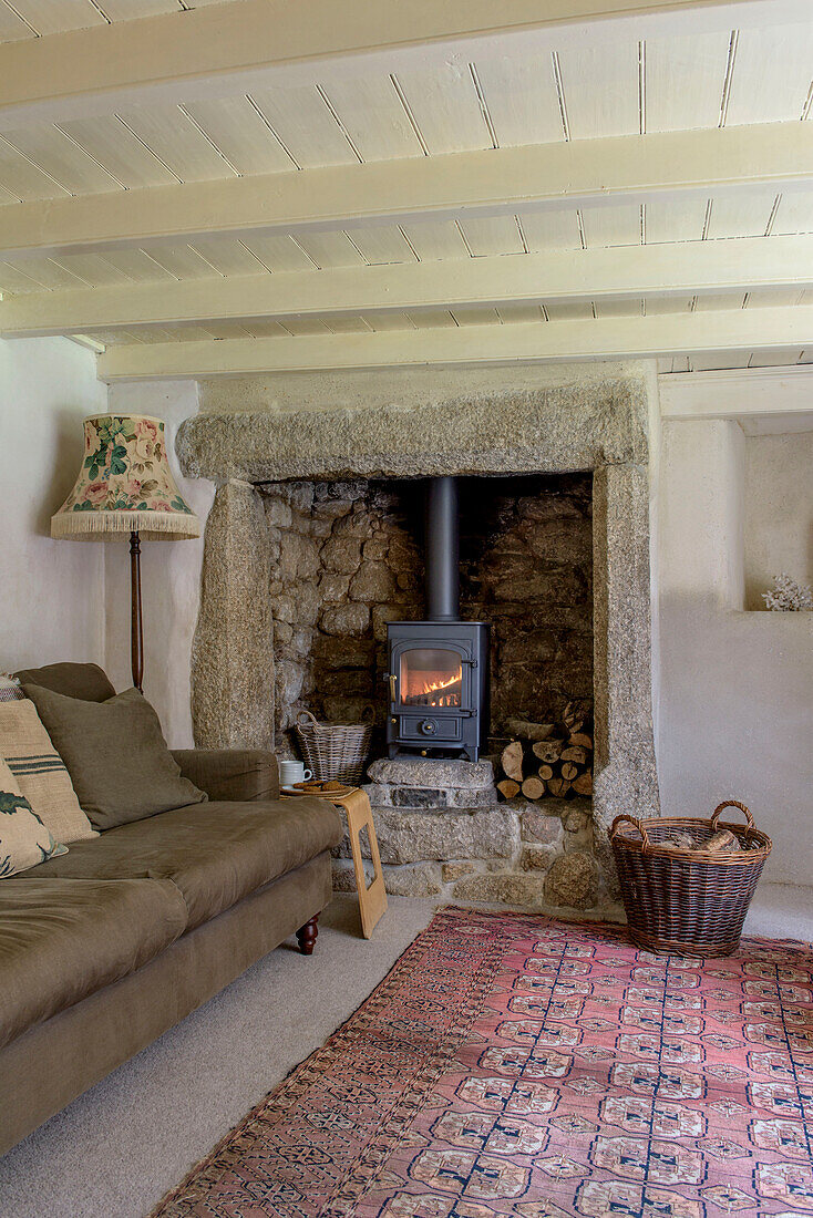 Braunes Sofa mit Stehlampe vor dem Kamin mit beleuchtetem Holzofen im Wohnzimmer eines Bauernhauses in Helston, Cornwall UK