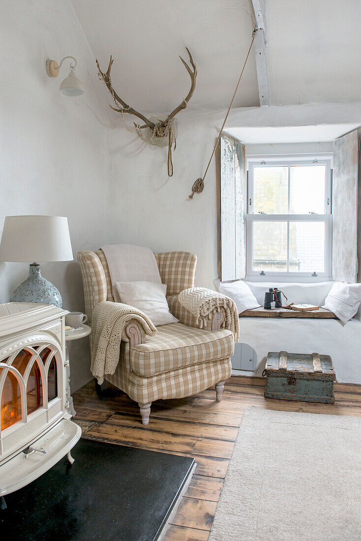 Gefaltete Decken auf kariertem Sessel mit beleuchtetem Holzofen im Wohnzimmer des Strandhauses in Marazion, Cornwall, UK