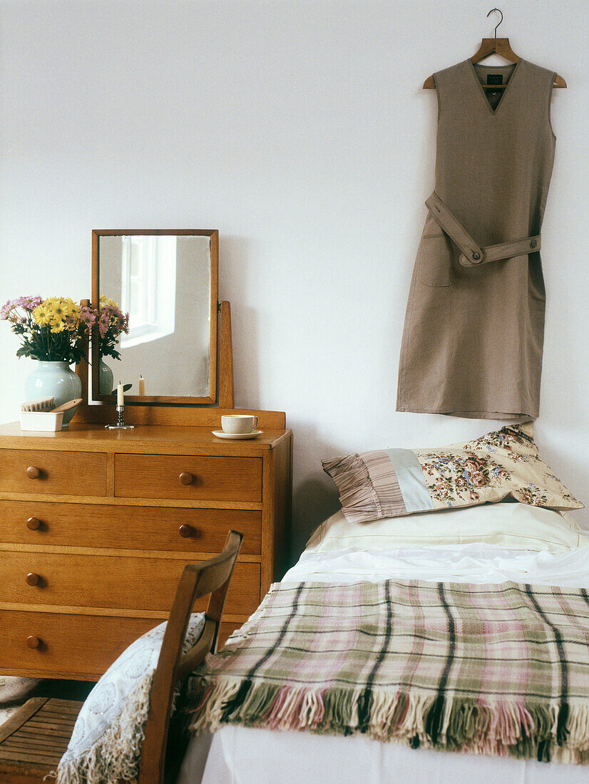 Utility style bedroom with wooden dressing table and mirror beside a single bed with blanket and 40s style dress on clotheshanger