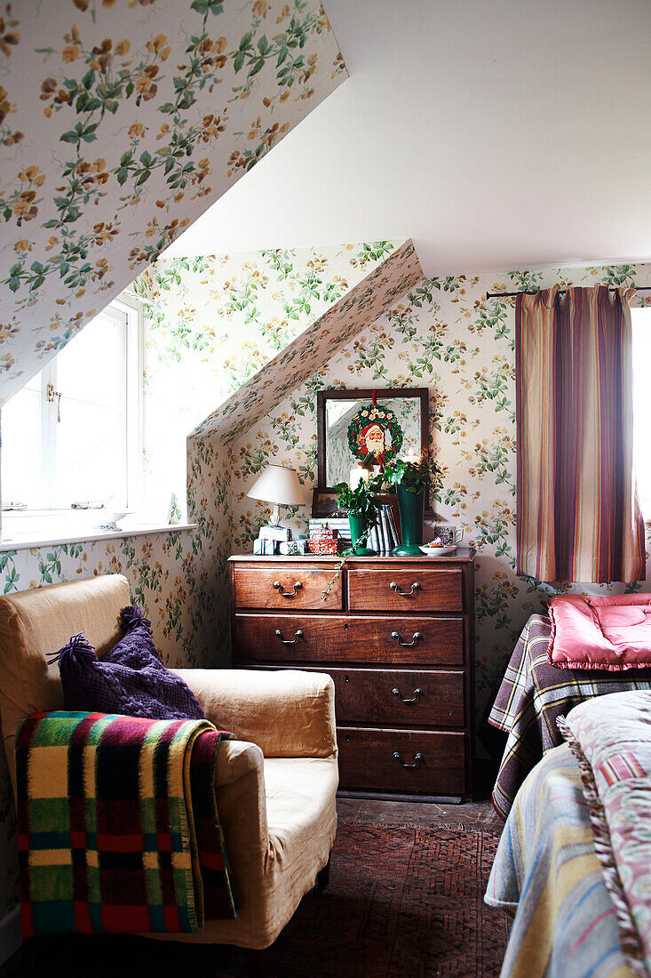 Sessel und Holzkommode unter der Dachgaube in einem mit Blumentapete tapezierten Schlafzimmer in einem Landhaus in Shropshire England UK