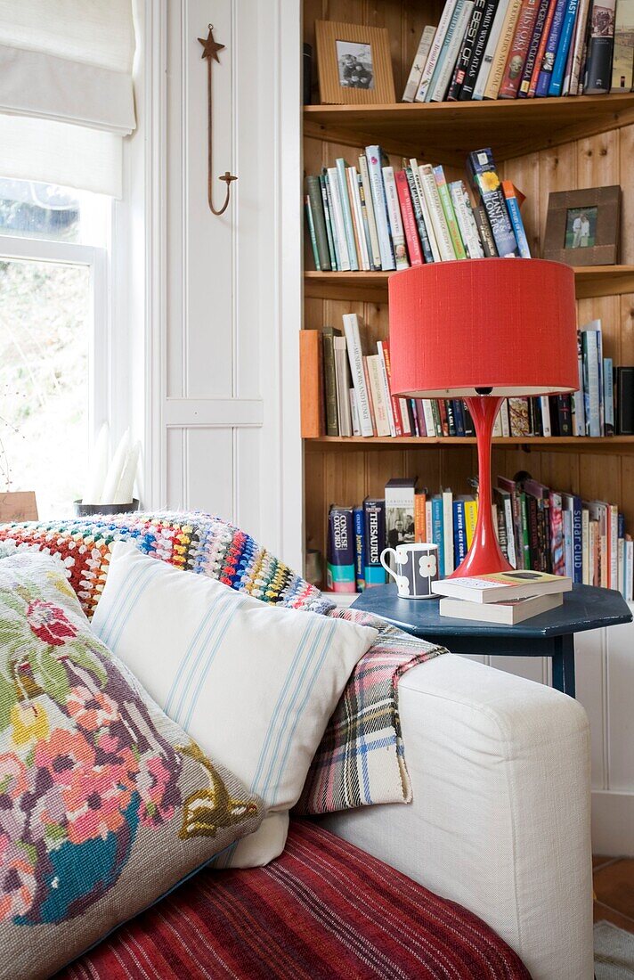 Rote Lampe auf Beistelltisch mit Bücherregal und Kissen im Haus einer Familie in Tenterden, Kent, England, Vereinigtes Königreich
