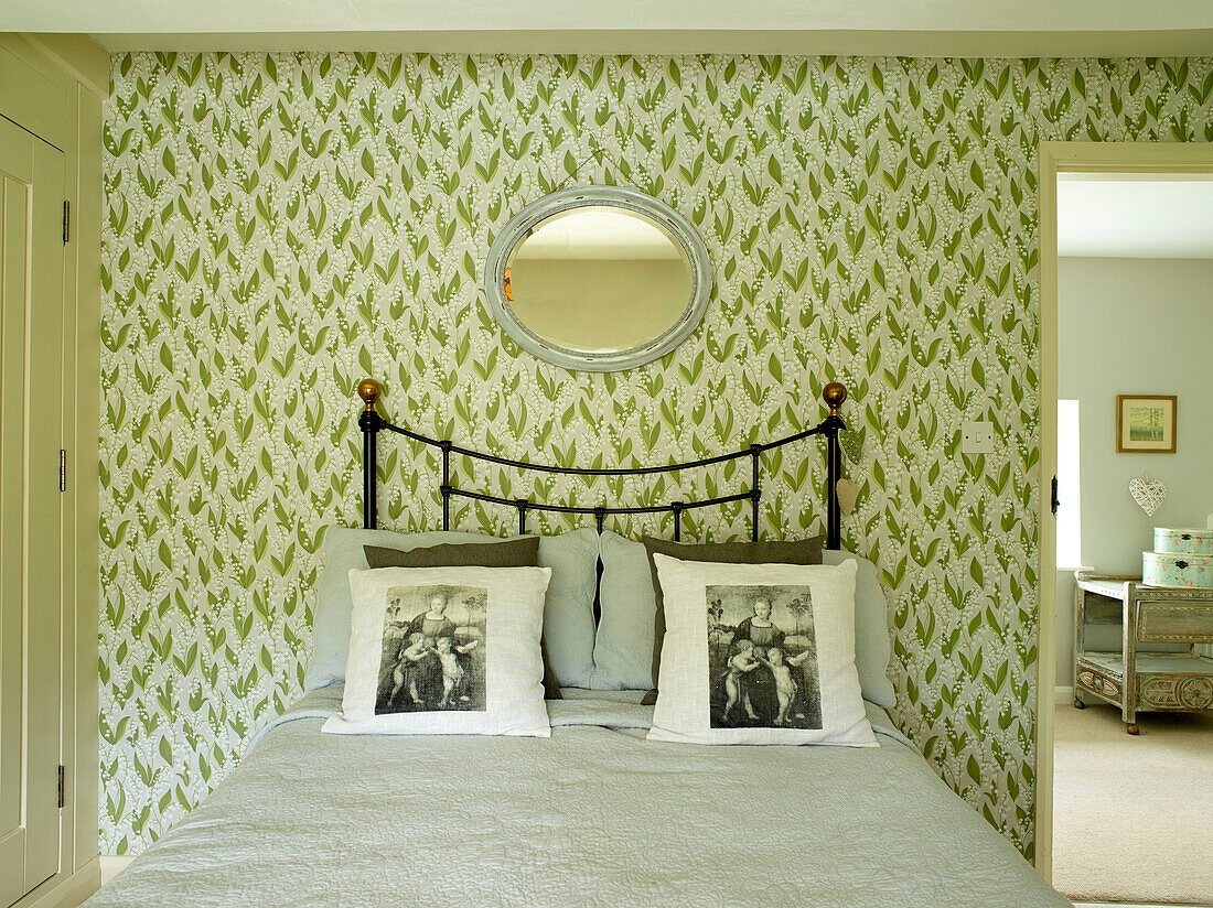 Ovaler Spiegel auf blattgemusterter Tapete über schmiedeeisernem Doppelbett in einem Landhaus in Oxfordshire, England, UK