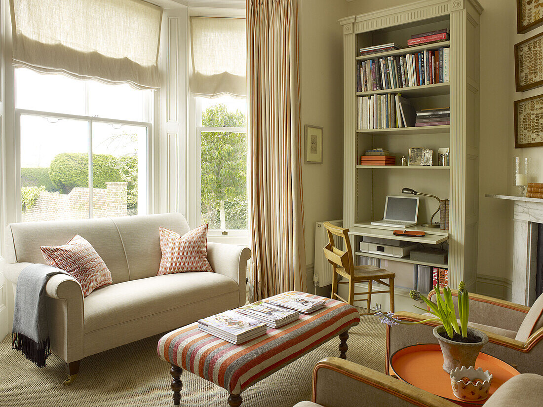 Sofa und gestreifter Ottomanenhocker mit Schreibtisch und Bücherregal am Fenster eines Landhauses in East Sussex (England)