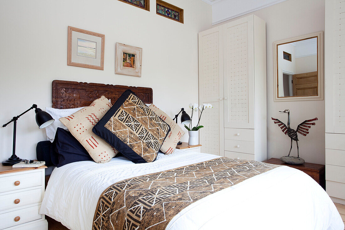 Patterned cushions on bed with carved headboard in white bedroom in London townhouse, England, UK