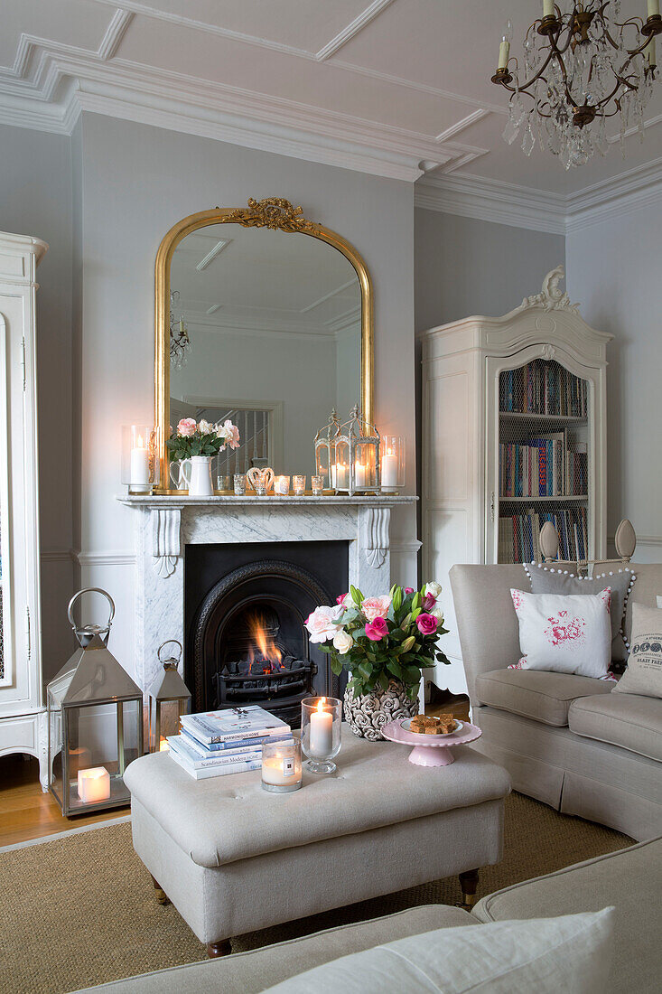 Lit candles with gilt framed mirror on mantlepiece with cut flowers on ottoman in living room of Hertfordshire home,  England,  UK